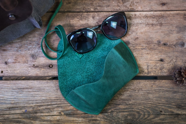 K&ouml;nnte beinhalten: Ein Sonnenbrillenetui aus tealgr&uuml;nem Leder mit einem Riemen. Das Etui liegt auf einer Holzoberfl&auml;che mit einer schwarzen Sonnenbrille darauf.
