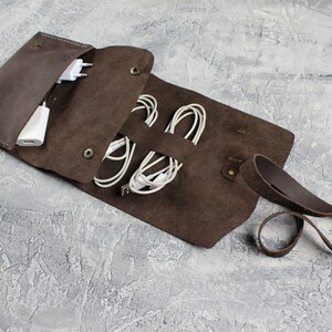 May include: A brown leather cord organizer with a flap closure and a strap. The organizer is open and contains white cords and a white adapter.