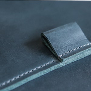 May include: Close-up of a dark teal leather item with a folded tab. The leather has a textured appearance and is stitched with white thread along the edges. The tab is folded over, creating a layered effect.