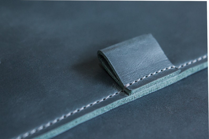 May include: Close-up of a dark teal leather item with a folded tab. The leather has a textured appearance and is stitched with white thread along the edges. The tab is folded over, creating a layered effect.