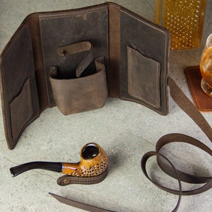 May include: A brown leather tobacco pouch with a strap and a pocket for a pipe. The pouch is open and a brown pipe is sitting next to it.