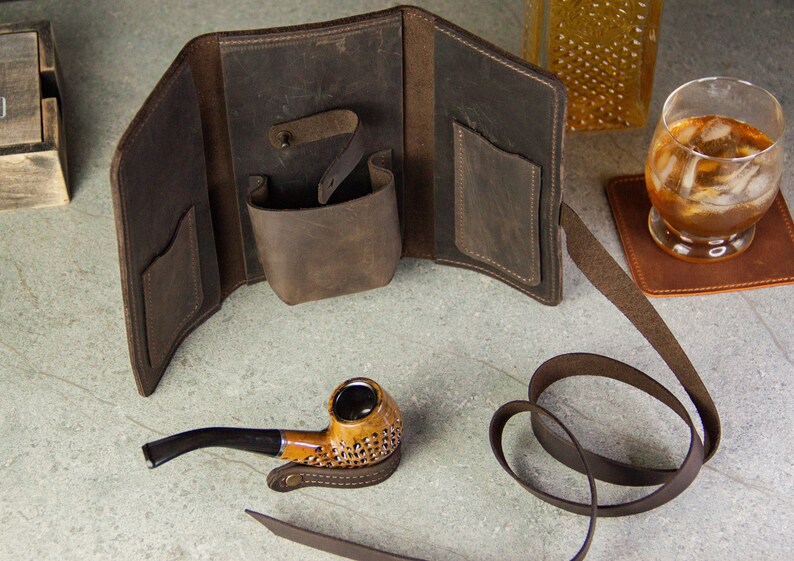 May include: A brown leather tobacco pouch with a strap and a pocket for a pipe. The pouch is open and a brown pipe is sitting next to it.