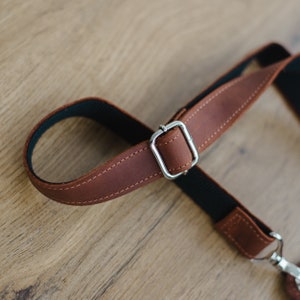 May include: Close-up of a brown leather camera strap with a silver metal buckle. The strap is adjustable and has a black lining.