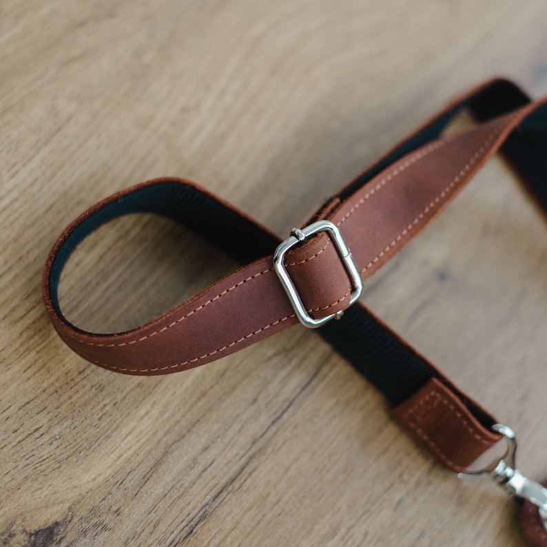 May include: Close-up of a brown leather camera strap with a silver metal buckle. The strap is adjustable and has a black lining.