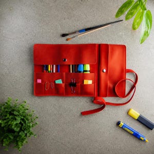 May include: A red leather roll-up organiser featuring multiple interior pockets filled with various art supplies, including coloured pencils, markers, paper clips, and sticky notes, secured with a matching leather strap and metal snap buttons.