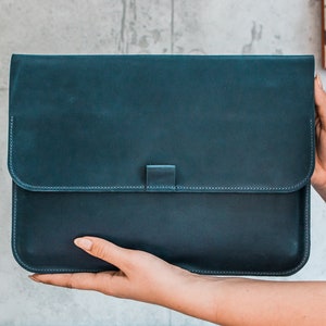 May include: A teal leather laptop sleeve with a flap closure. The sleeve is rectangular with rounded corners and a stitched border. The flap is secured with a small tab. The leather has a slightly distressed appearance.