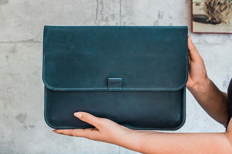 May include: A teal leather laptop sleeve with a flap closure. The sleeve is rectangular with rounded corners and a stitched border. The flap is secured with a small tab. The leather has a slightly distressed appearance.