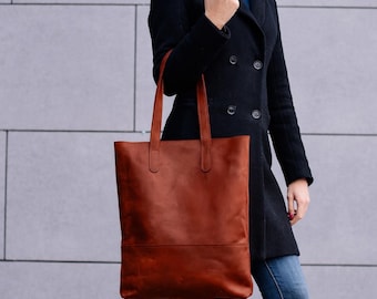 May include: A brown leather tote bag with long handles. The bag is rectangular with a simple design and a lighter brown section at the bottom. The bag is being held in front of a person wearing a black coat and blue jeans.