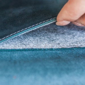 May include: Close-up of a teal leather item with a gray felt interior. The leather has a textured appearance and is stitched with white thread. The felt interior is visible as the item is opened.
