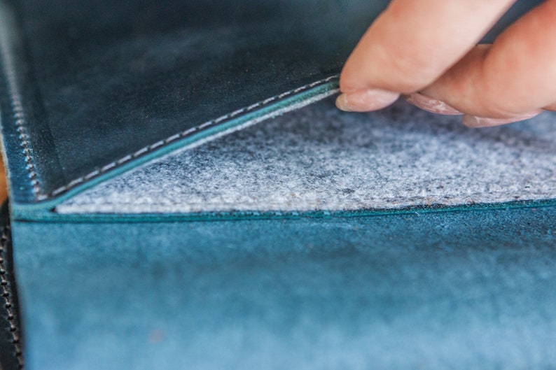 May include: Close-up of a teal leather item with a gray felt interior. The leather has a textured appearance and is stitched with white thread. The felt interior is visible as the item is opened.