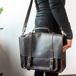 May include: A dark brown leather briefcase with a long strap and two buckles. The briefcase has a top handle and is worn over the shoulder.