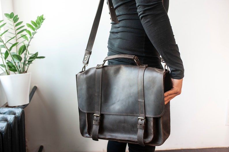 May include: A dark brown leather briefcase with a long strap and two buckles. The briefcase has a top handle and is worn over the shoulder.