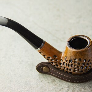 May include: A brown wooden smoking pipe with a black stem and a leather carrying case. The pipe has a decorative pattern on the bowl.