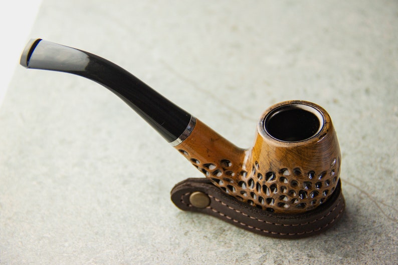 May include: A brown wooden smoking pipe with a black stem and a leather carrying case. The pipe has a decorative pattern on the bowl.