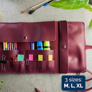 May include: A burgundy leather roll-up organiser featuring multiple interior pockets and slots filled with coloured pencils, markers, paper clips, and sticky notes. The organiser includes a snap closure and a text graphic that reads 3 sizes: M, L, XL.
