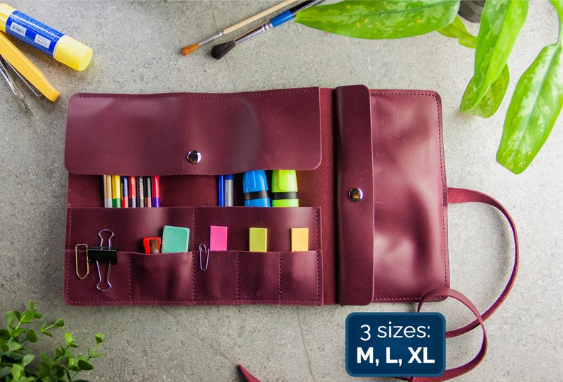 May include: A burgundy leather roll-up organiser featuring multiple interior pockets and slots filled with coloured pencils, markers, paper clips, and sticky notes. The organiser includes a snap closure and a text graphic that reads 3 sizes: M, L, XL.