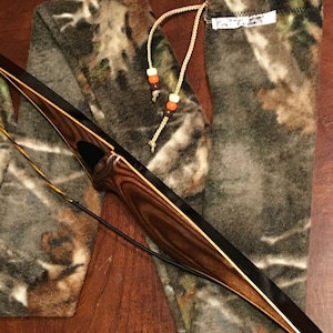 May include: A camouflage bow case with a drawstring closure and a brown leather strap. The case is made of a soft, fleece-like material and has a brown and white camouflage pattern.