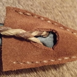 May include: A brown leather sheath with a hole for a blade, stitched with brown thread. A braided rope is threaded through the hole.