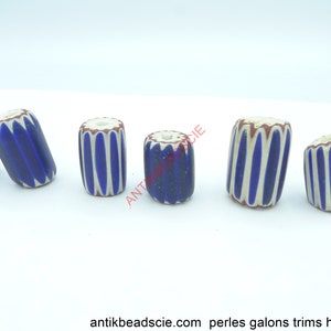 May include: Six antique glass beads with a cylindrical shape. Each bead features a blue and white striped pattern, with a brown and white rim. The beads are displayed on a white surface. The text "antikbeadscie.com" is visible at the bottom.