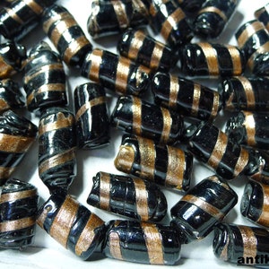 Könnte beinhalten: Eine Ansammlung schwarzer Glasperlen mit goldfarbenen Streifen. Die zylindrischen Perlen haben eine glänzende Oberfläche. Die Perlen sind verstreut, und das Bild ist gut beleuchtet.