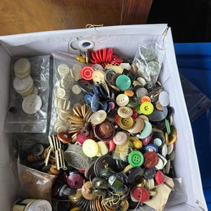 Peut inclure: Une boîte débordant d'une collection variée de boutons de différentes tailles, formes et couleurs. L'assortiment comprend des boutons blancs, noirs, rouges, verts et bruns, certains dans de petits sacs en plastique. Une petite boîte avec une illustration de pingouin est également visible.