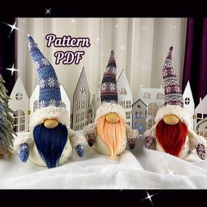 May include: Three handmade gnome ornaments with white fur, red, blue, and pink beards, and patterned knit hats. The gnomes are sitting on a white surface with a wooden background.