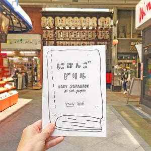 Puede incluir: Un folleto de papel blanco con texto negro e ilustraciones. El texto dice "Easy Japanese for cool people" y "Study Book". El folleto está siendo sostenido por una mano en un fondo borroso.