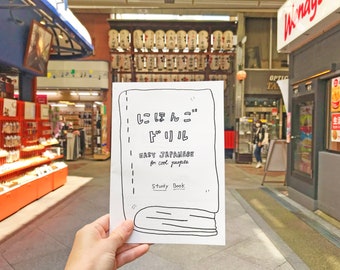 May include: A white paper booklet with black text and illustrations. The text reads "Easy Japanese for cool people" and "Study Book". The booklet is being held by a hand in a blurry background.