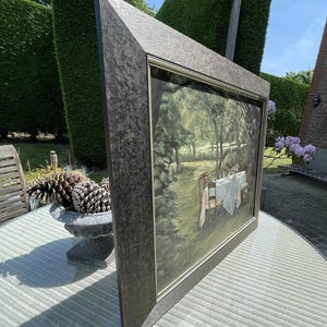 May include: A framed painting depicting a table set for a meal in a garden setting. The painting has a dark, textured frame. The scene includes a white tablecloth, chairs, and lush greenery. The painting is displayed outdoors.