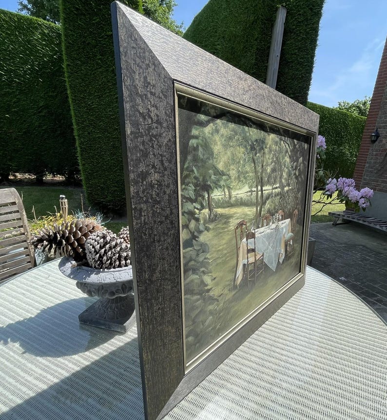 May include: A framed painting depicting a table set for a meal in a garden setting. The painting has a dark, textured frame. The scene includes a white tablecloth, chairs, and lush greenery. The painting is displayed outdoors.