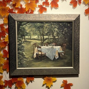 May include: A framed painting depicts a serene outdoor dining scene. A table is set with a white tablecloth, surrounded by wooden chairs. The artwork is framed in a dark, textured frame, and surrounded by decorative autumn leaves.