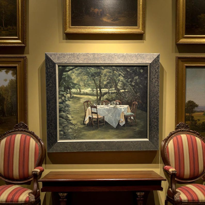 May include: An oil painting of a table set for a meal in a garden, surrounded by trees. The painting is in a dark gray frame and is displayed on a wall with other framed paintings. Two chairs with red and cream striped upholstery are in the foreground.