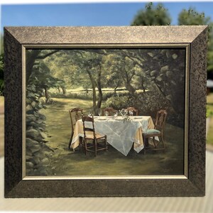 May include: A framed painting depicts a serene outdoor dining scene. A white tablecloth covers a table set with glasses and flowers. Four wooden chairs surround the table. The painting is set in a lush garden with trees and a stone wall.