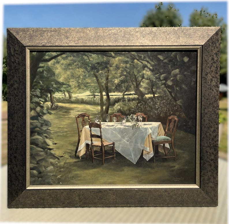 May include: A framed painting depicts a serene outdoor dining scene. A white tablecloth covers a table set with glasses and flowers. Four wooden chairs surround the table. The painting is set in a lush garden with trees and a stone wall.