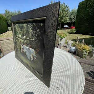 May include: A framed painting depicting a dining table set for a meal in a garden setting. The painting has a dark, textured frame. The table is covered with a white tablecloth and set with chairs. The scene is bathed in natural light.
