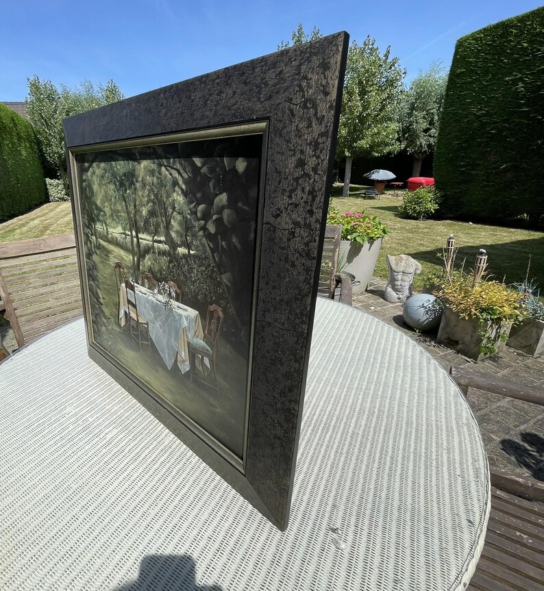 May include: A framed painting depicting a dining table set for a meal in a garden setting. The painting has a dark, textured frame. The table is covered with a white tablecloth and set with chairs. The scene is bathed in natural light.