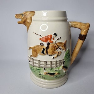 May include: Cream-colored ceramic pitcher with a hunting scene. The spout is shaped like a fox head. A raised relief depicts a rider on a horse jumping over a fence, with dogs in pursuit. The handle is brown and tan.