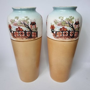 May include: A pair of tall, ceramic vases with a light blue and tan glaze. The vases feature a hand-painted design of a landscape with trees, houses, and a fence.