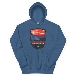 May include: A blue hooded sweatshirt featuring a graphic print of a sunset over a landscape. The text reads EXPLORE KENTUCKY, INDIAN STAIRCASE, RED RIVER GORGE, and L48. The design is shaped like a shield with a red sky, orange sun, and blue hills.