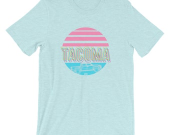 May include: A light blue t-shirt featuring a pink and blue sunset graphic. The graphic includes the word "TACOMA" in a rainbow font and a silhouette of a pickup truck.