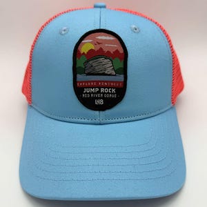 May include: A light blue trucker hat with a coral mesh back and a curved brim. The front features a patch with a landscape design and the text "EXPLORE KENTUCKY JUMP ROCK - RED RIVER GORGE - L48".