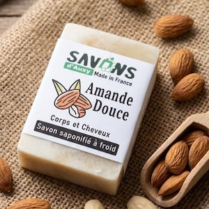 Könnte beinhalten: Ein rechteckiges Stück Seife, umwickelt mit einem weißen Papieretikett mit dem Text Savons d'Auxy Made in France, Amande Douce, Corps et Cheveux und Savon saponifié à froid, verziert mit einer Mandelillustration und verstreuten Mandeln.