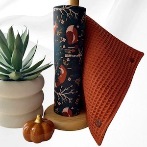 Könnte beinhalten: Ein Papierrollenhalter aus Holz mit einer Stoffrollenbespannung in Dunkelblau, bedruckt mit orangefarbenen Füchsen, weißen Blumen und Bäumen. Ein rostfarbenes, waffelstrukturiertes Stofftuch mit braunen Druckknöpfen hängt an der Rolle.