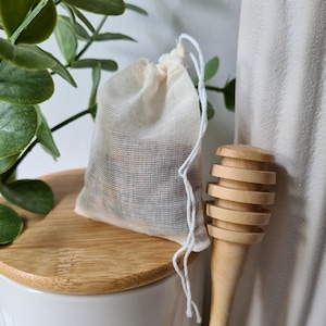 Könnte beinhalten: Ein kleiner, cremefarbener Beutel mit Kordelzug aus gewebtem Stoff, der dunklen Inhalt durch das Material erkennen lässt. Neben dem Beutel befindet sich ein heller Honiglöffel aus Holz mit gerilltem Kopf und langem, glattem Stiel.