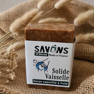 Könnte beinhalten: Ein rechteckiges, braun gesprenkeltes Spülmittelstück, umwickelt mit einem weißen Papieretikett. Das Etikett zeigt den Text SAVONS d'Auxy Made in France, eine Grafik einer Hand, die einen blauen Teller wäscht, und den Text Solide Vaisselle Savon saponifié à froid.
