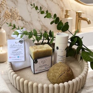 Peut inclure: Un plateau beige texturé contient un shampoing solide, un pain de savon, un bâton de déodorant et un savon rond. Le savon est étiqueté "Savon Mojito". Une branche de feuilles vertes et un sac blanc avec une étiquette "Bulle de Lagon" sont également présents.