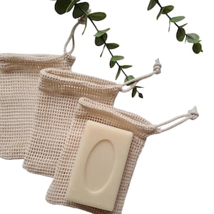 Peut inclure: Trois sacs à savon en filet beige avec fermetures à cordon, dont un contenant un pain de savon de couleur claire. Des brins de feuilles vertes sont en arrière-plan. Les sacs sont en matériau tissé.