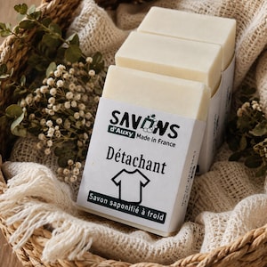 Könnte beinhalten: Drei rechteckige, cremefarbene Seifenstücke, die in weiße Papieretiketten eingewickelt sind. Die Etiketten tragen den Text SAVONS d'Auxy Made in France, das Wort Détachant, ein T-Shirt-Symbol und den Text Savon saponifié à froid.