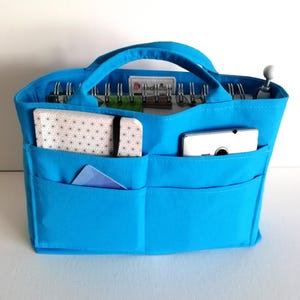 Può includere: Un organizer per borsa in tessuto blu brillante con due manici superiori e molteplici tasche esterne. L'interno contiene un quaderno a spirale, mentre le tasche frontali ospitano un quaderno bianco con motivo geometrico, uno smartphone bianco e una carta.
