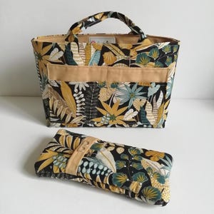 Puede incluir: Un bolso tote y un clutch a juego con un estampado botánico negro, amarillo, verde y blanco. El bolso tote tiene un bolsillo y asas color beige. El clutch tiene una correa beige con cierre de botón.